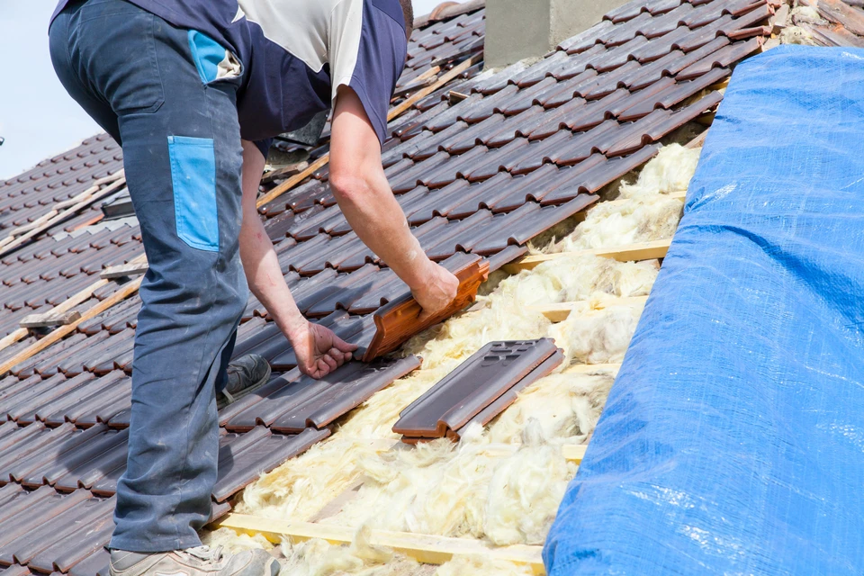 Photographie d'un couvreur expérimenté en tenue de travail de sécurité (gilet haute visibilité, casque) travaillant sur une toiture d'une maison résidentielle. L'ouvrier est photographié en action, posant des tuiles ou effectuant des travaux de zinguerie sur la pente d'un toit, démontrant clairement son expertise technique et son engagement sur le chantier. La maison en arrière-plan est de style résidentiel classique français. La photographie est prise de jour, avec une lumière naturelle claire mettant en évidence la qualité du travail et le professionnalisme de l'intervention.