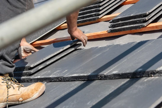 Photographie en gros plan d'un int&eacute;rieur de grenier ou comble montrant une trace d'infiltration d'eau ou une tache d'humidit&eacute; sombre sur la face inf&eacute;rieure des chevrons ou sur la couche d'isolation, avec une goutte d'eau ou des gouttes visibles. La lumi&egrave;re naturelle provient d'une fen&ecirc;tre ou d'une ouverture de toit. L'image doit montrer clairement les d&eacute;g&acirc;ts caus&eacute;s par une fuite (coulure, aur&eacute;ole sombre) et transmettre l'urgence d'une r&eacute;paration toiture professionnelle. Photo de jour, nette, professionnelle, sans texte ni logo.